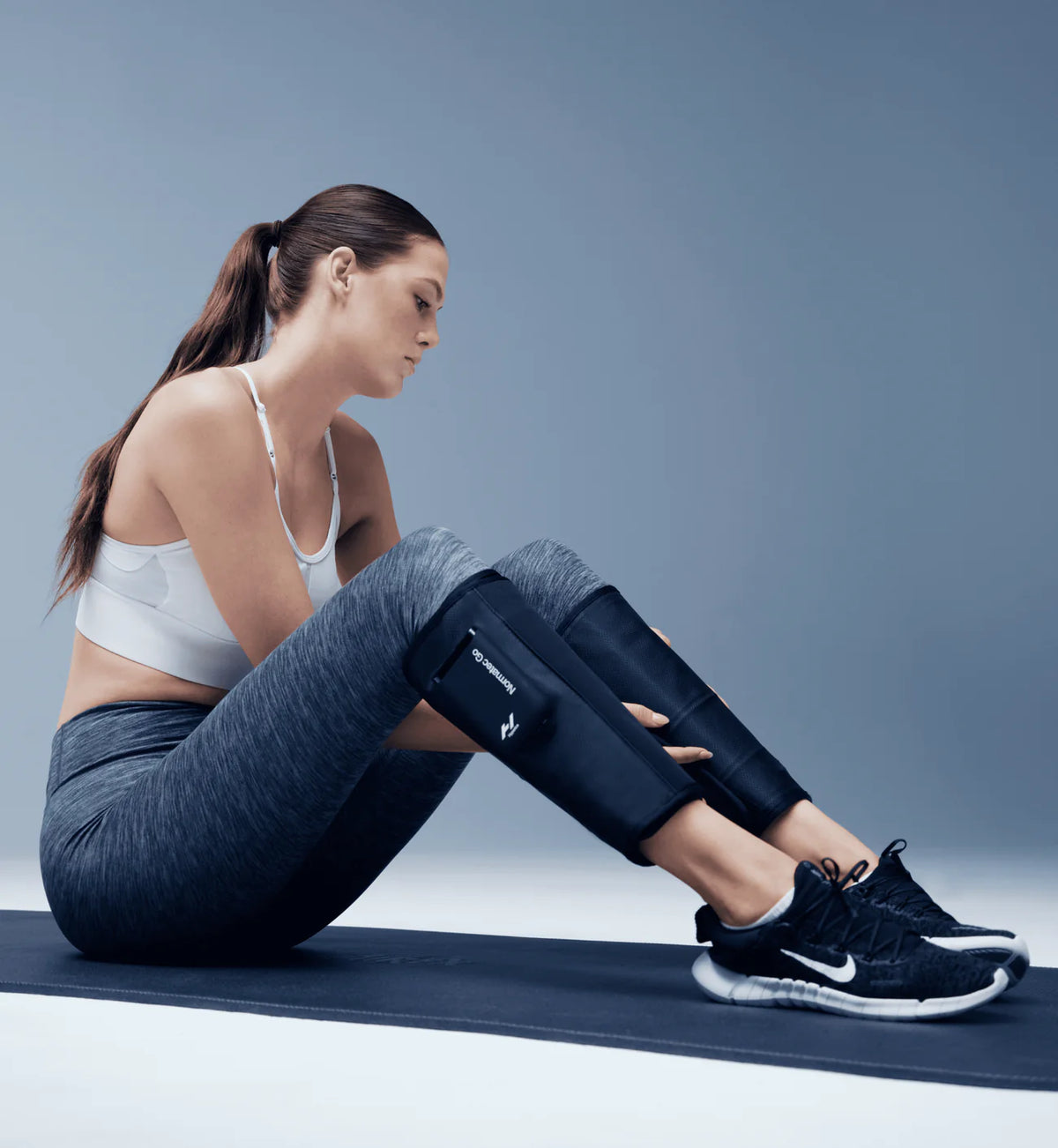 Woman sitting on a yoga mat wearing Hyperice Normatec Go compression sleeves on calves